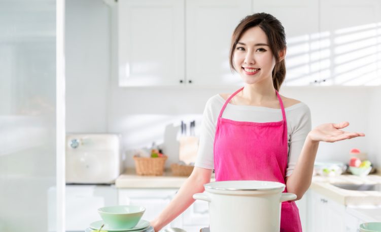 キッチンで火のついた鍋の前で微笑みながら左手を横に差し出している若い女性