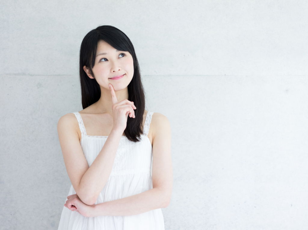 右手の人差し指を顎に当てて上向き目線で考え事をしている白いワンピースの女性