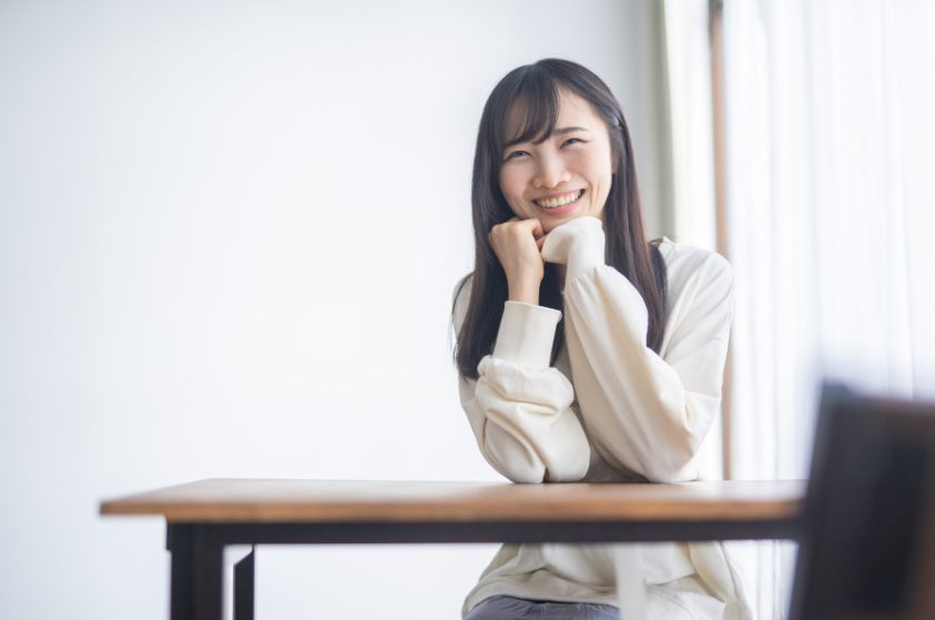 テーブルの前に座り、両拳を顎に当て嬉しそうに微笑む白いセーターを着た若い女性