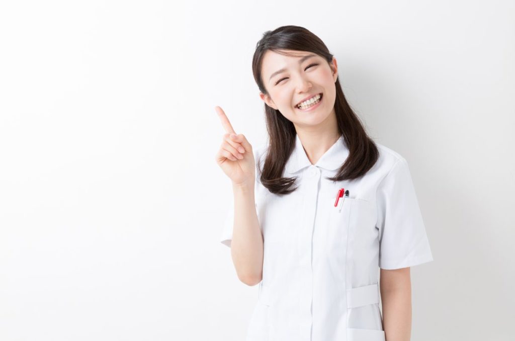 白い医療服を着て、右人差し指を差し出す笑顔の若い女性