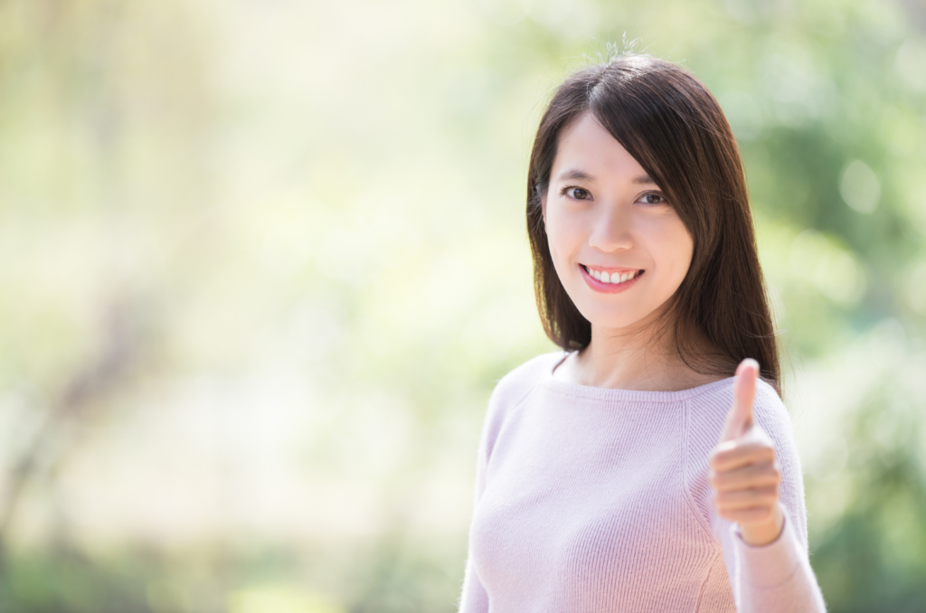 ピンクのセーターを着た、左親指を突き出して微笑んでいる薄い若い女性