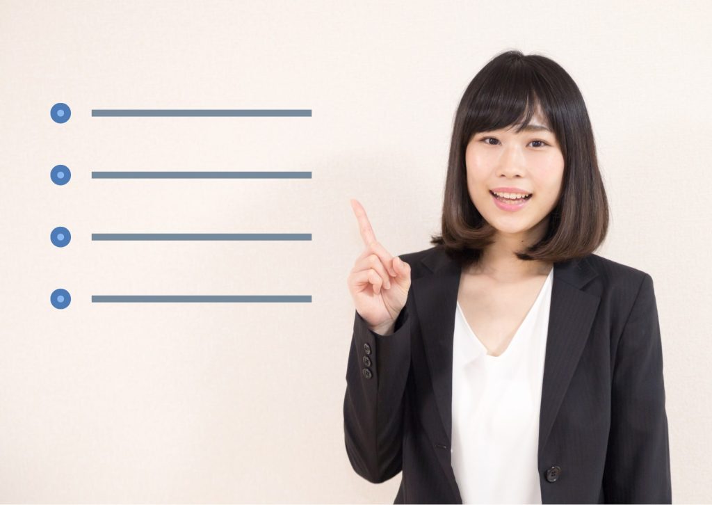 背景の段落を右人差し指指で指し、笑顔でしゃべる若い黒スーツの女性