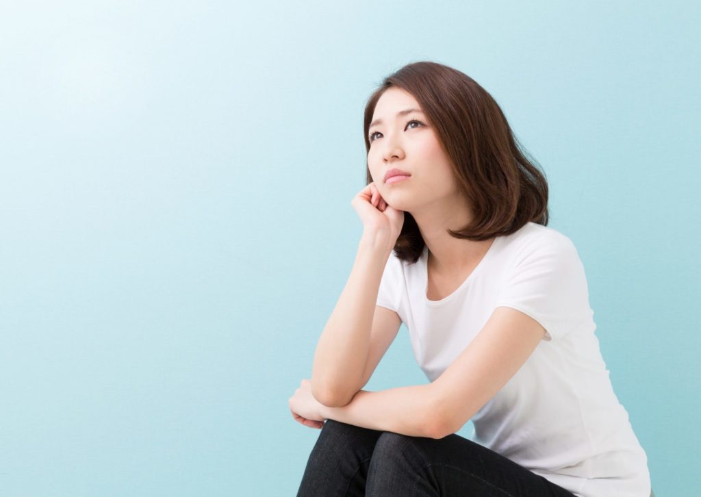 水色の背景で、若い女性が顎に右券を当てて上を見て考え事をしている