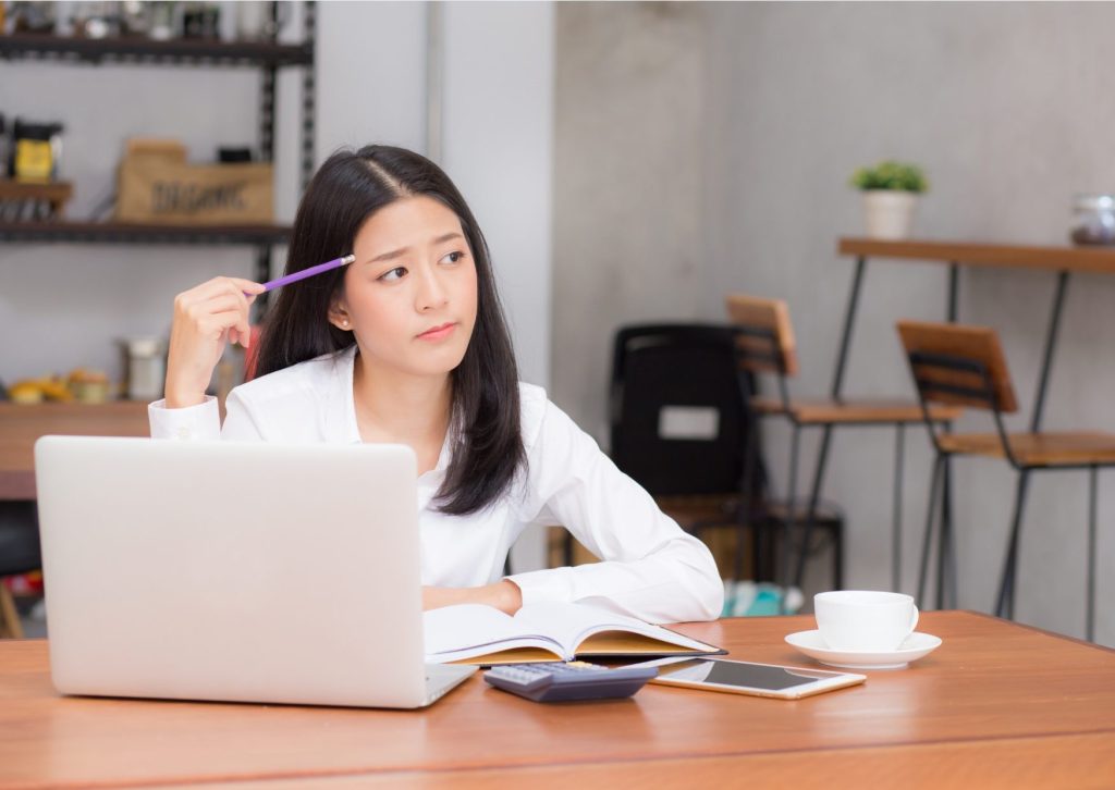 机の上のノートパソコンの前に座り、シャーペンをおでこに当てて、考え込む白シャツの若い女性