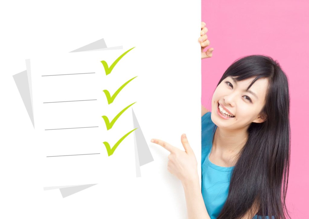 ボードのレ点リストを指さし、ボード後ろから上半身をのぞかせるブルーのワンピースの若い女性