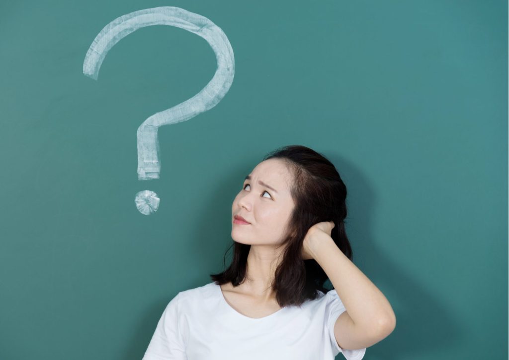 左手を頭の後ろに当て、斜め上を向いて困り顔で考え事をしている若い女性の上に？マーク