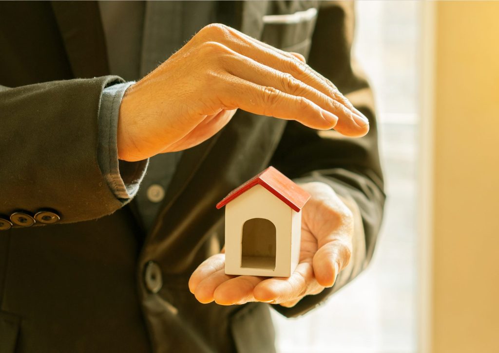 スーツの男性が左手の平に家のおもちゃを乗せ、右手で屋根の形をしておもちゃの屋根の上でかざしてる