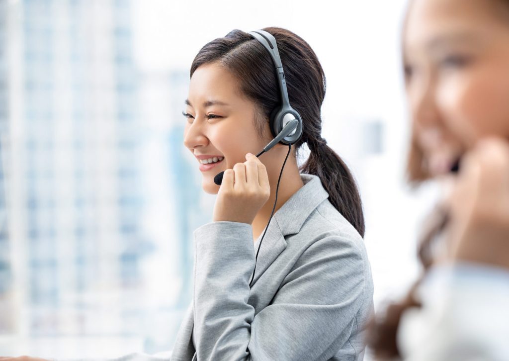 インカムを付けたグレースーツを着た笑顔の若い女性。左手でマイクを支えている