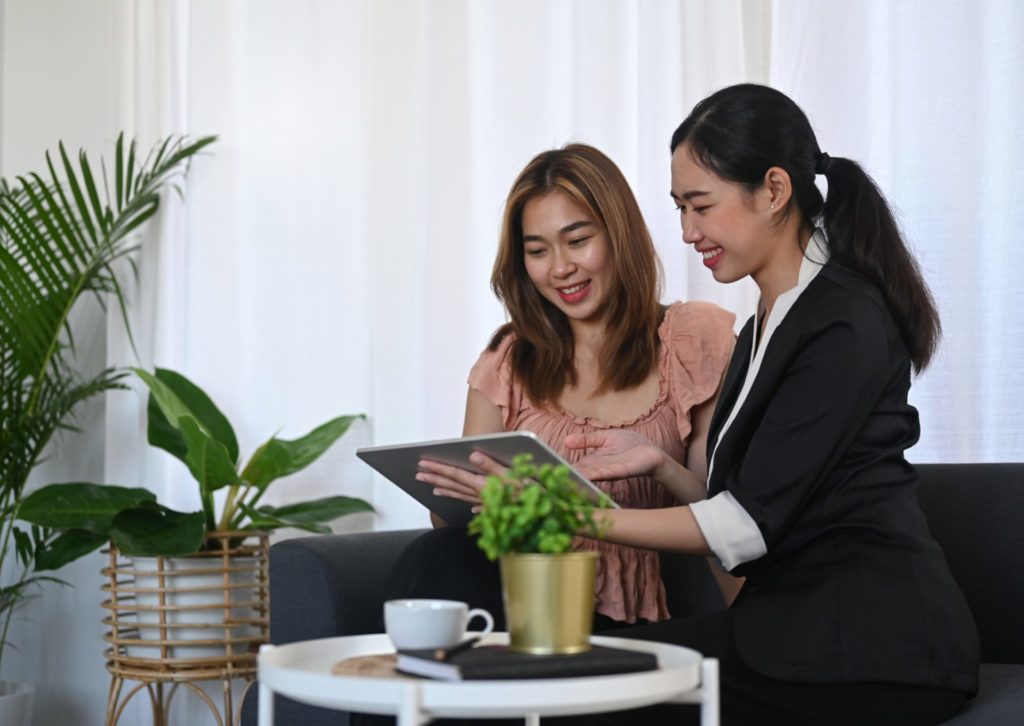 黒いスーツの若い女性がタブレットでピンクのワンピースの若い女性に説明をしている