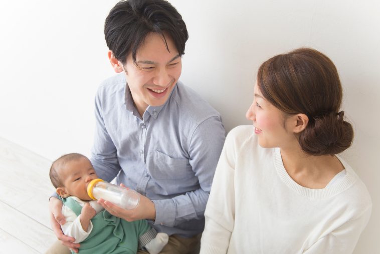 パパが赤ちゃんにミルクを飲ませている、ママがその横で微笑んでお互いの科をお見つめあっている